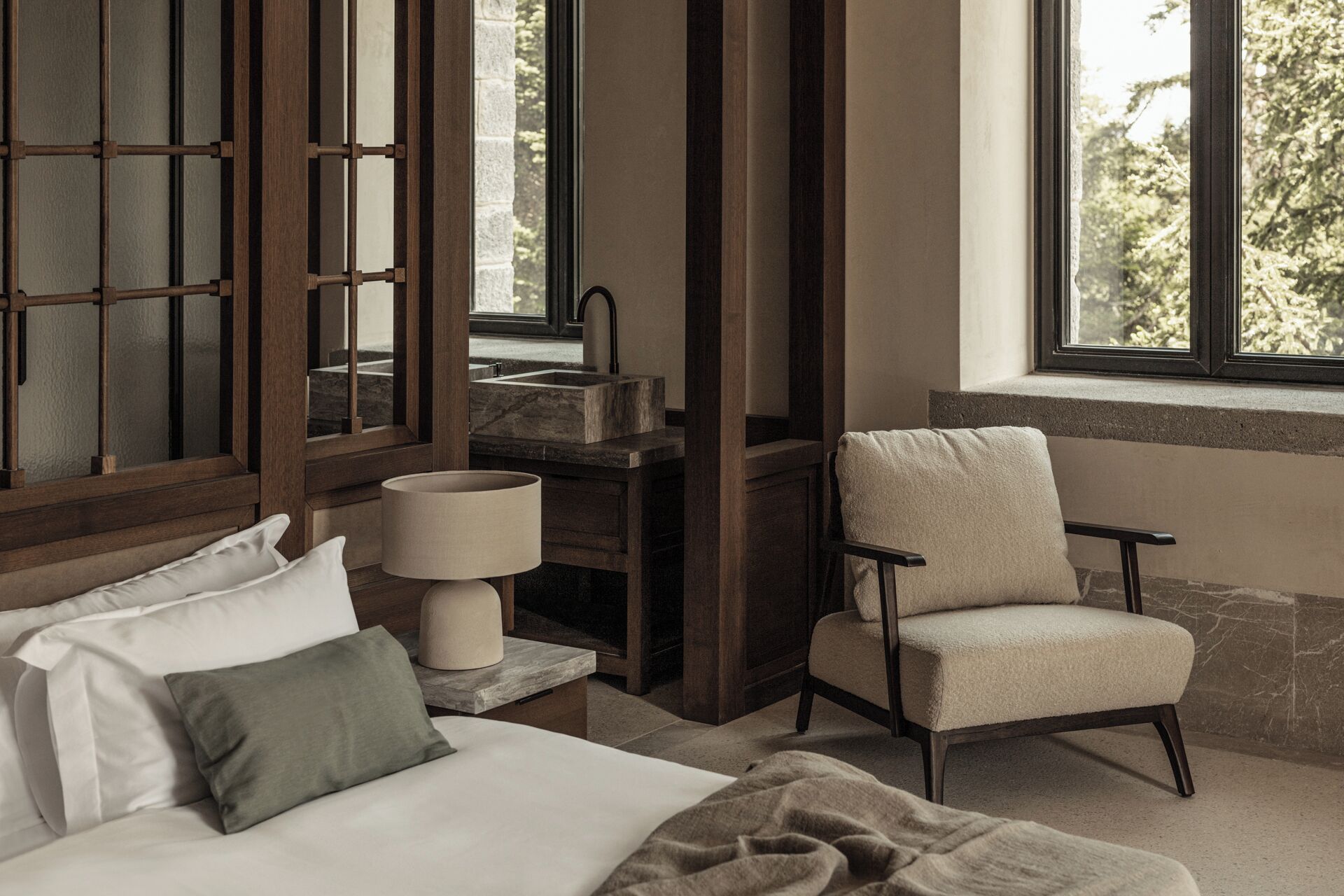 A bedroom with a bed, a chair, a nightstand, a lamp, and a window with a view of trees.