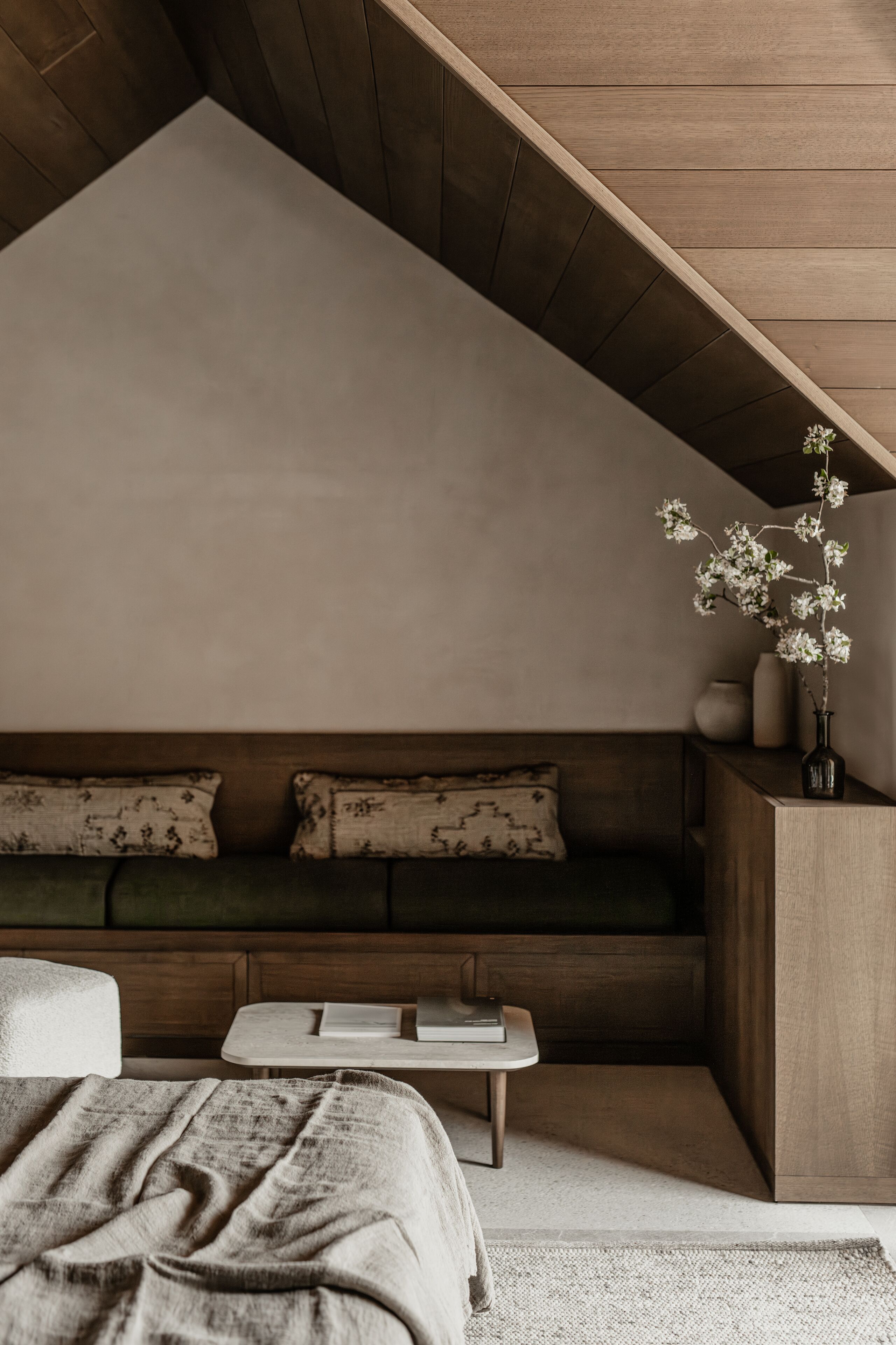 Quarto moderno com teto inclinado, um banco de madeira, mesa de cabeceira e um vaso com flores.