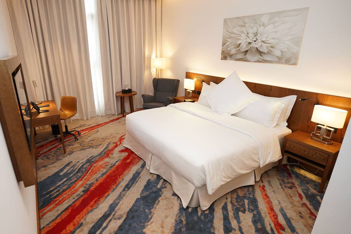 A hotel room with a large bed, a desk with a chair, a small table, and a lamp.