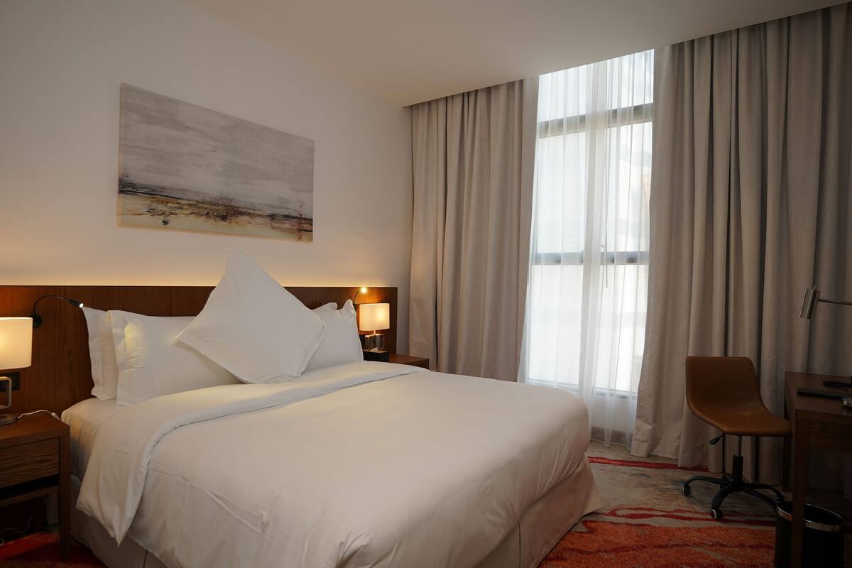 A hotel room with a large bed, two bedside tables with lamps, a desk with a chair, a large window with curtains, and a painting on the wall.
