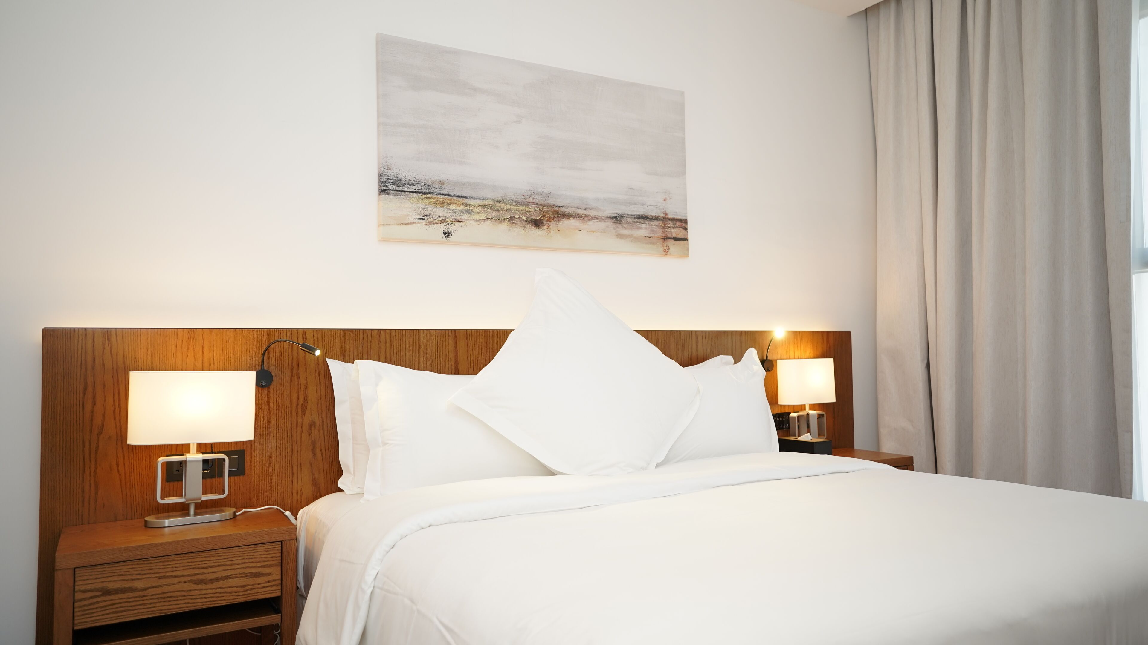 Habitación de hotel con una cama grande, cabecera de madera, dos mesitas de noche con lámparas, un cuadro en la pared y vistas a la playa a través de la ventana.