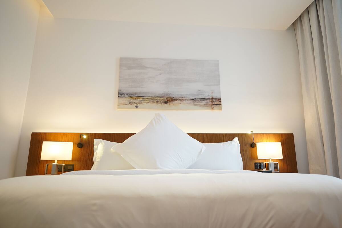 A hotel room with a large bed, two bedside lamps, a painting on the wall, and a window with curtains.