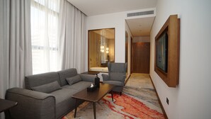 Habitación de hotel moderna con un sofá gris, una mesita pequeña, un sillón gris y un espejo grande que refleja un baño con bañera.
