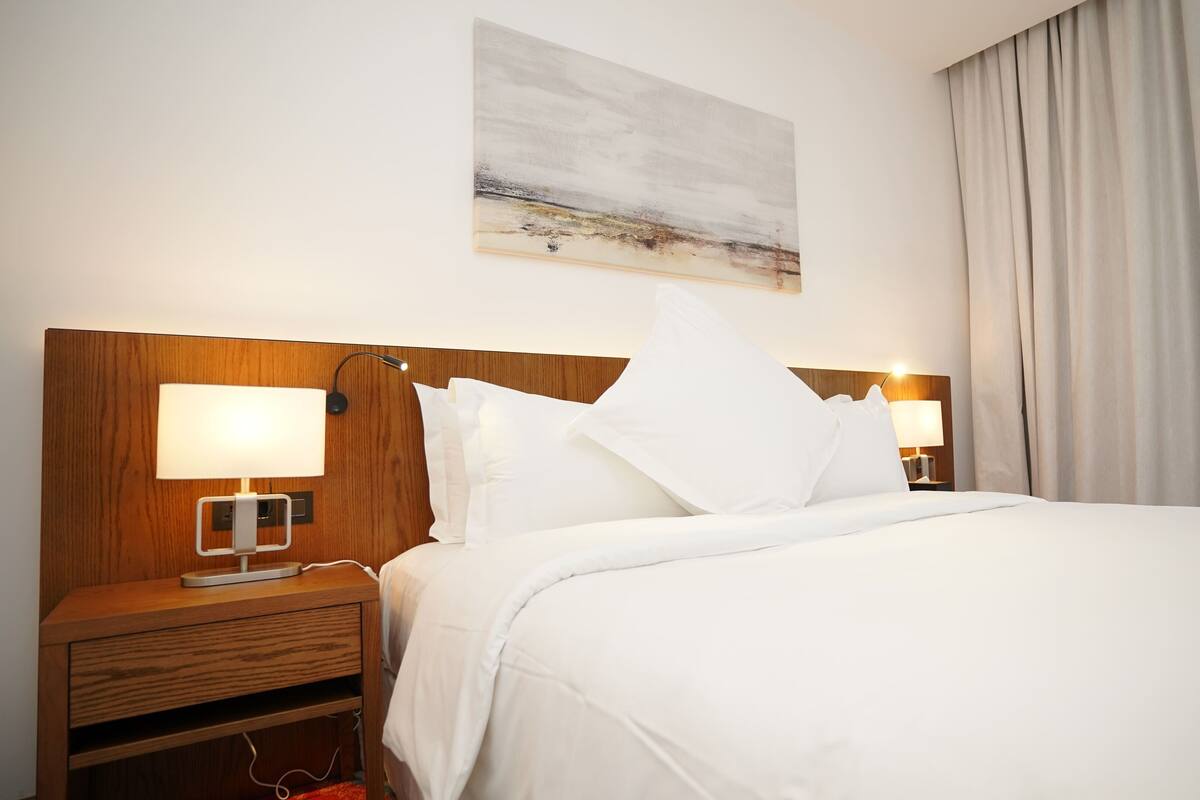 A hotel room with a wooden headboard, two bedside lamps, a large bed with white bedding, a painting on the wall, and a window with curtains.