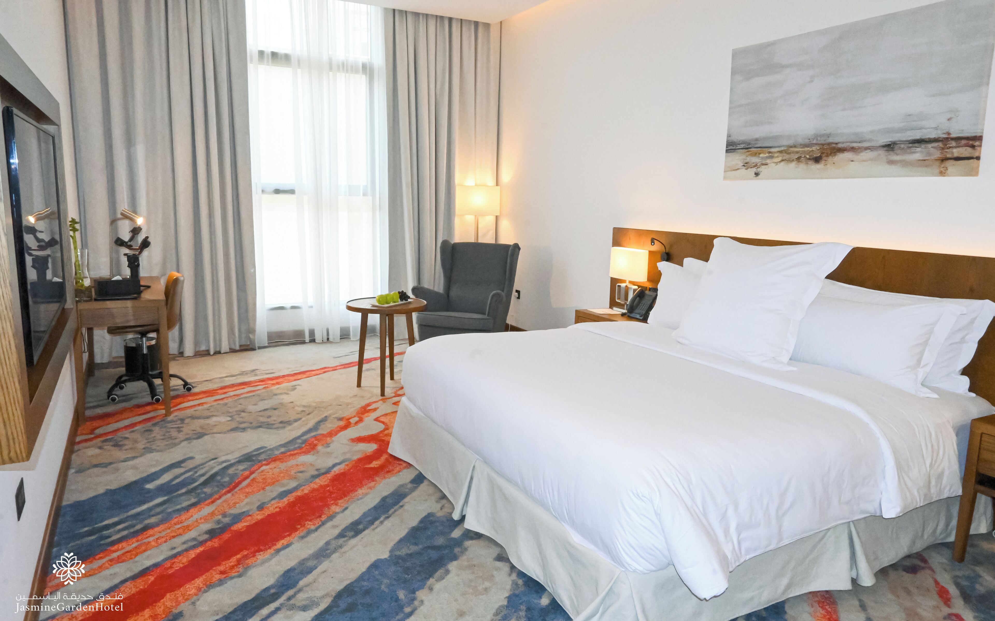 Une chambre d’hôtel avec un grand lit, un bureau avec une chaise, une petite table avec des fruits, une lampe et un tableau au mur.