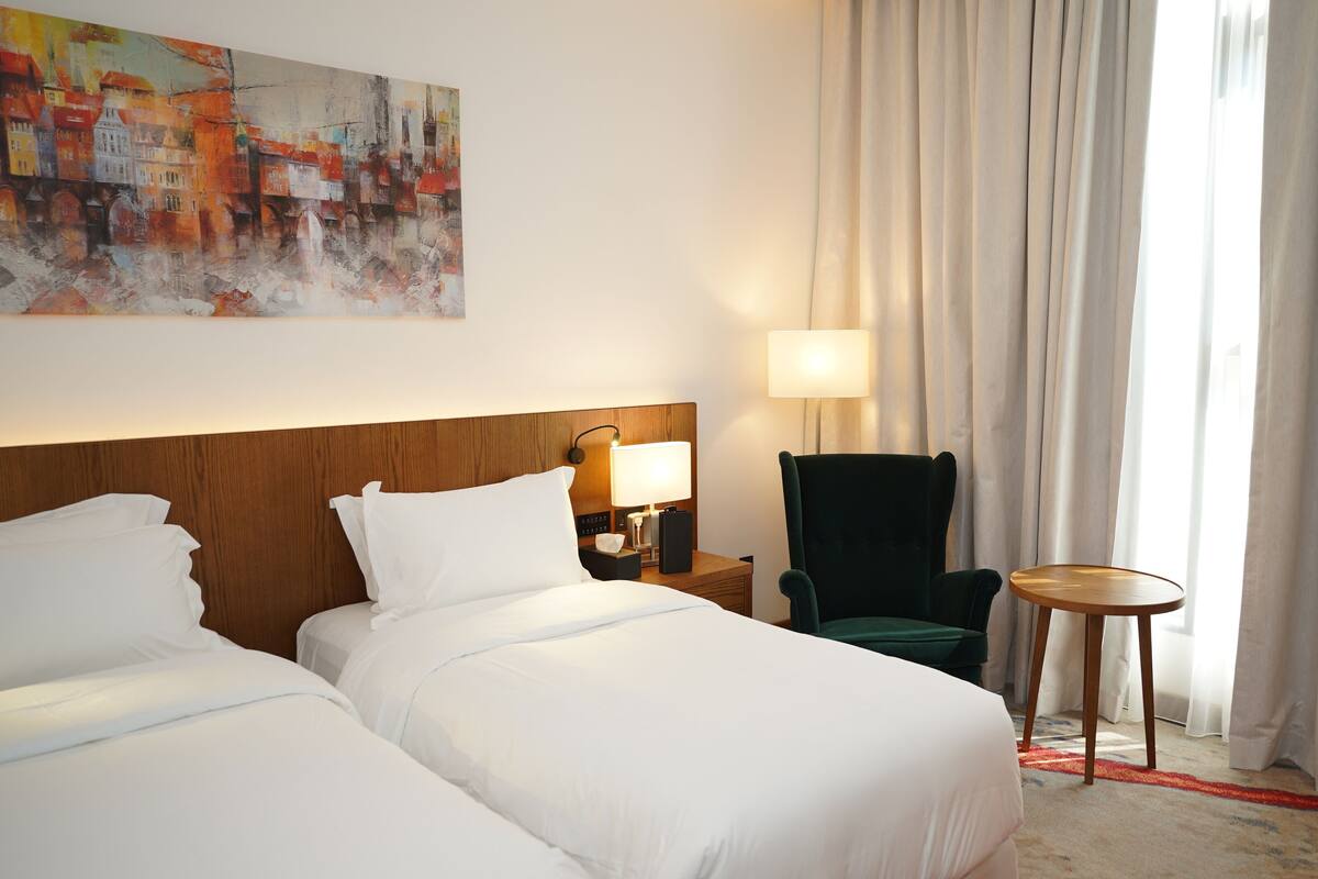 A hotel room with two beds, a chair, a small table, a lamp, and a painting on the wall.