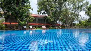 A swimming pool with clear blue water, surrounded by lush greenery and a building with a red roof. - Oleander Farms (Khalapur)