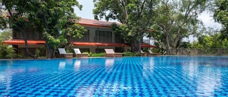 A swimming pool with clear blue water, surrounded by lush greenery and a building with a red roof.