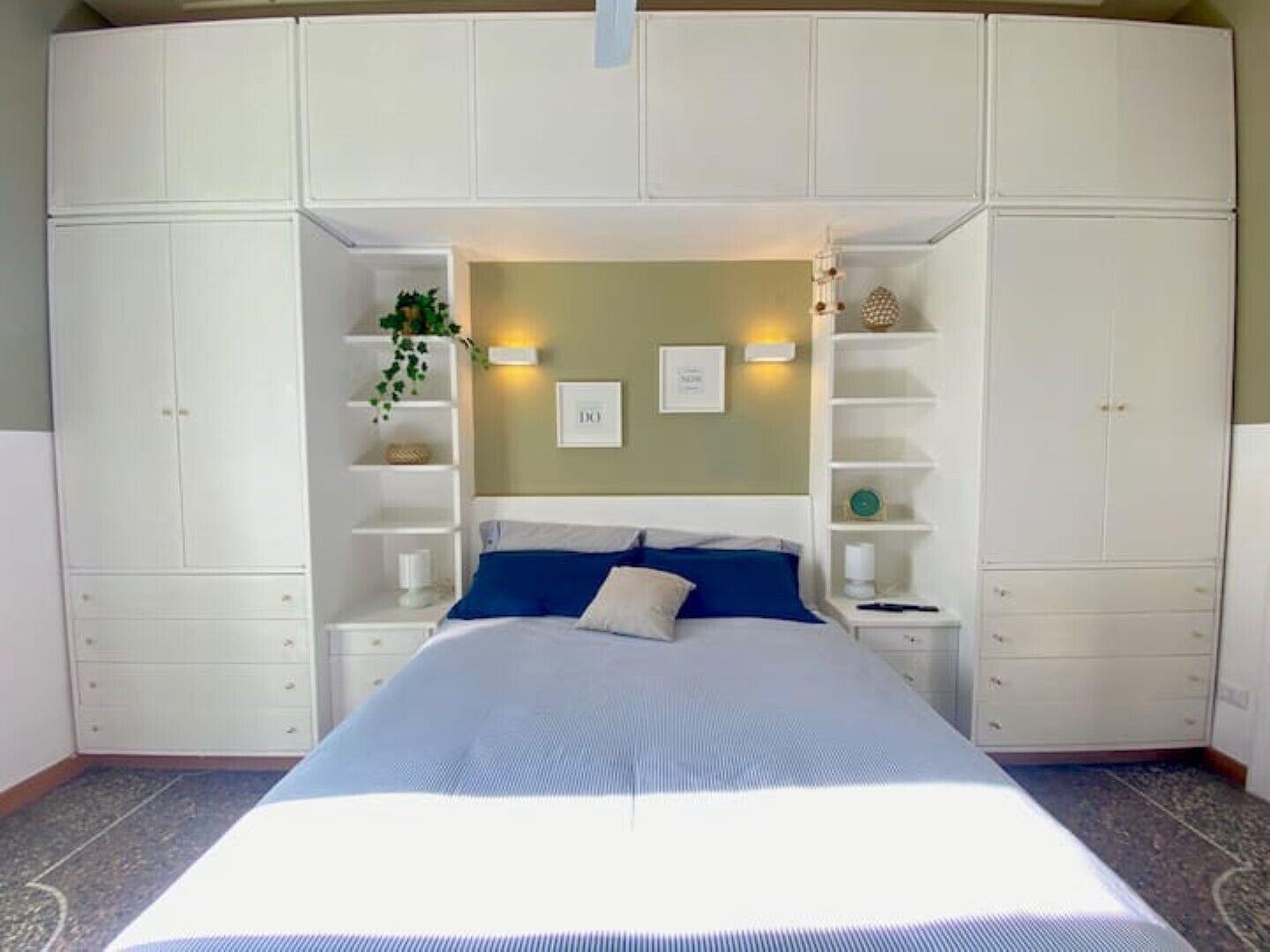 Una camera da letto con un letto al centro, sotto un armadio bianco con ripiani e cassetti, due luci a parete e una piccola pianta a sinistra.