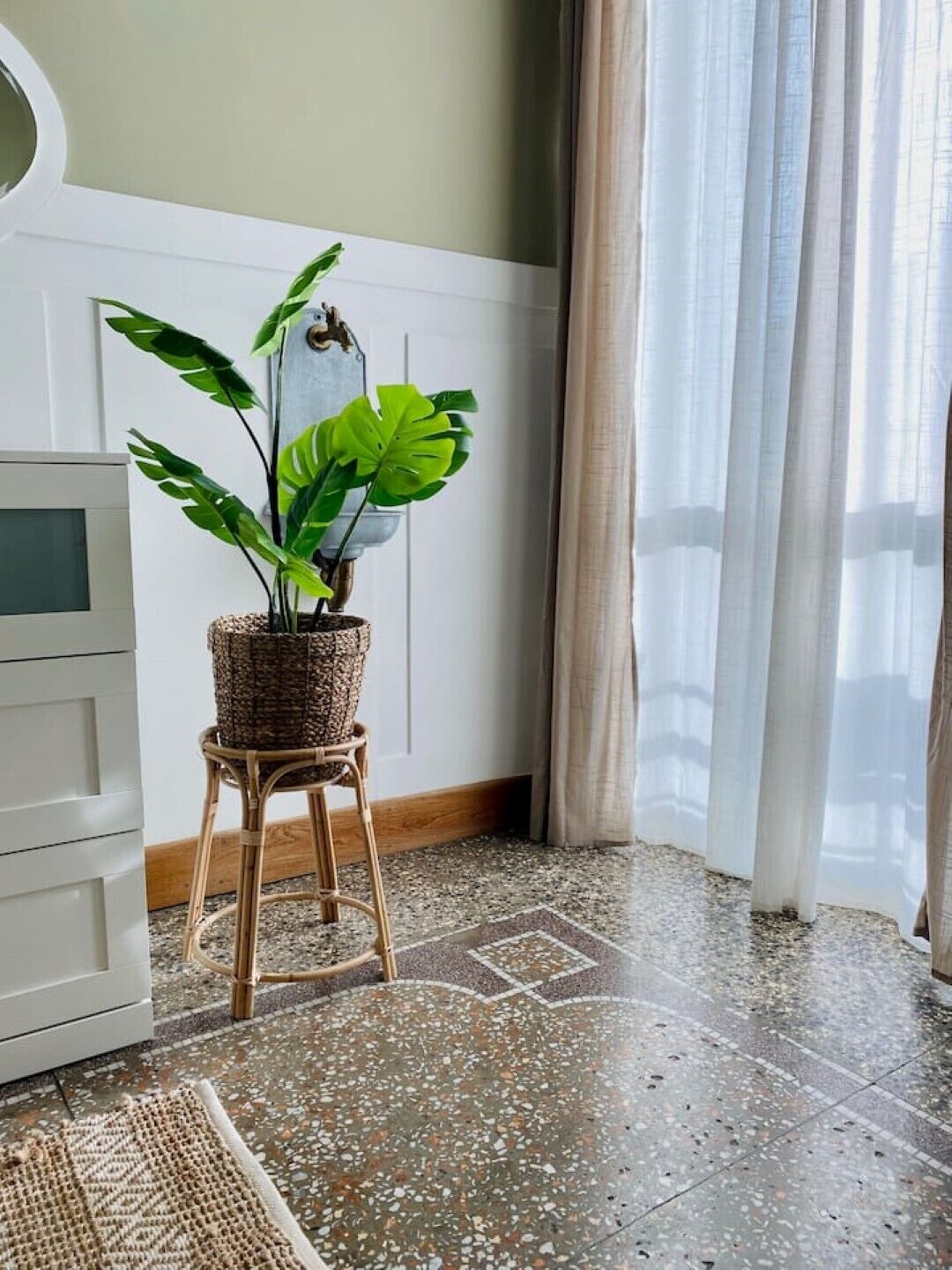Una habitación con una planta en maceta sobre un taburete, un armario blanco y una ventana con cortinas.