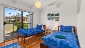 A bedroom with two beds, a balcony, and a view of buildings and greenery. - Baxter's Beach House by Experience Jervis Bay (Vincentia)