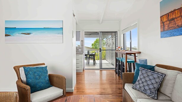 A modern living room with wicker furniture, a glass door leading to a balcony, and a painting of a beach scene. - Baxter's Beach House by Experience Jervis Bay (Vincentia)