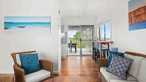 Una sala de estar moderna con mobiliario de mimbre, un ventanal de vidrio que da a un balcón y un cuadro con una escena de playa.