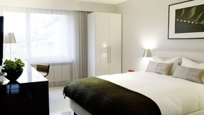 Una habitación de hotel moderna con una cama grande, un escritorio con lámpara, una silla y una ventana con cortinas.
