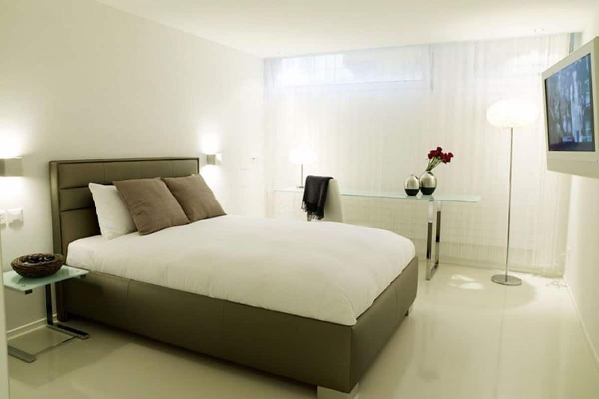 Une chambre moderne avec un grand lit, des tables de chevet, un bureau en verre et une lampe sur pied.