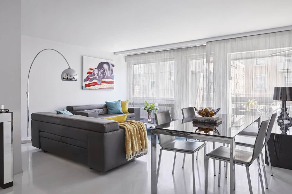 Un salon moderne avec un canapé gris, une table à manger avec des chaises et une grande fenêtre ornée de rideaux légers.