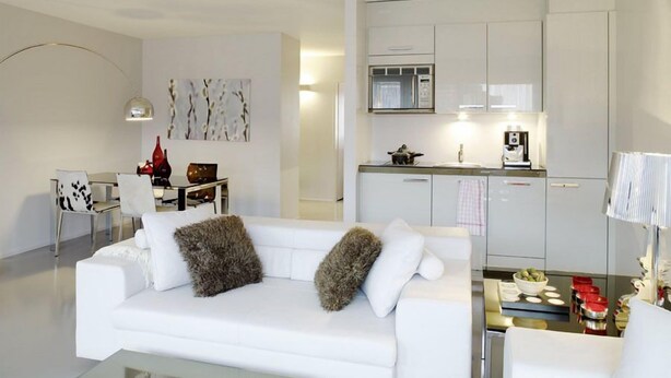 Una sala de estar moderna con un sofá blanco, una mesa de centro y una cocina con armarios y electrodomésticos blancos.