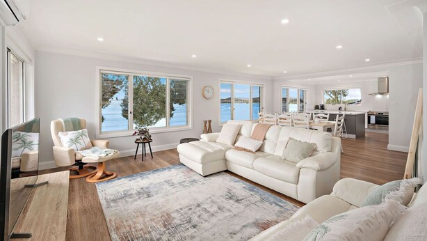 Ein geräumiges Wohnzimmer mit einem großen, weißen Ecksofa, einem Couchtisch und einem Fernseher. Große Fenster bieten Blick auf das Meer.