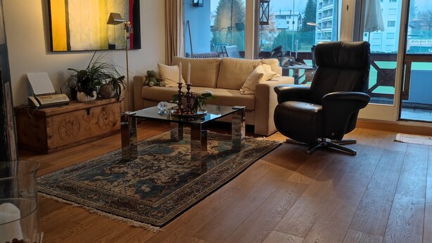 A spacious living room with a large window, a sofa, a black leather chair, and a wooden coffee table.