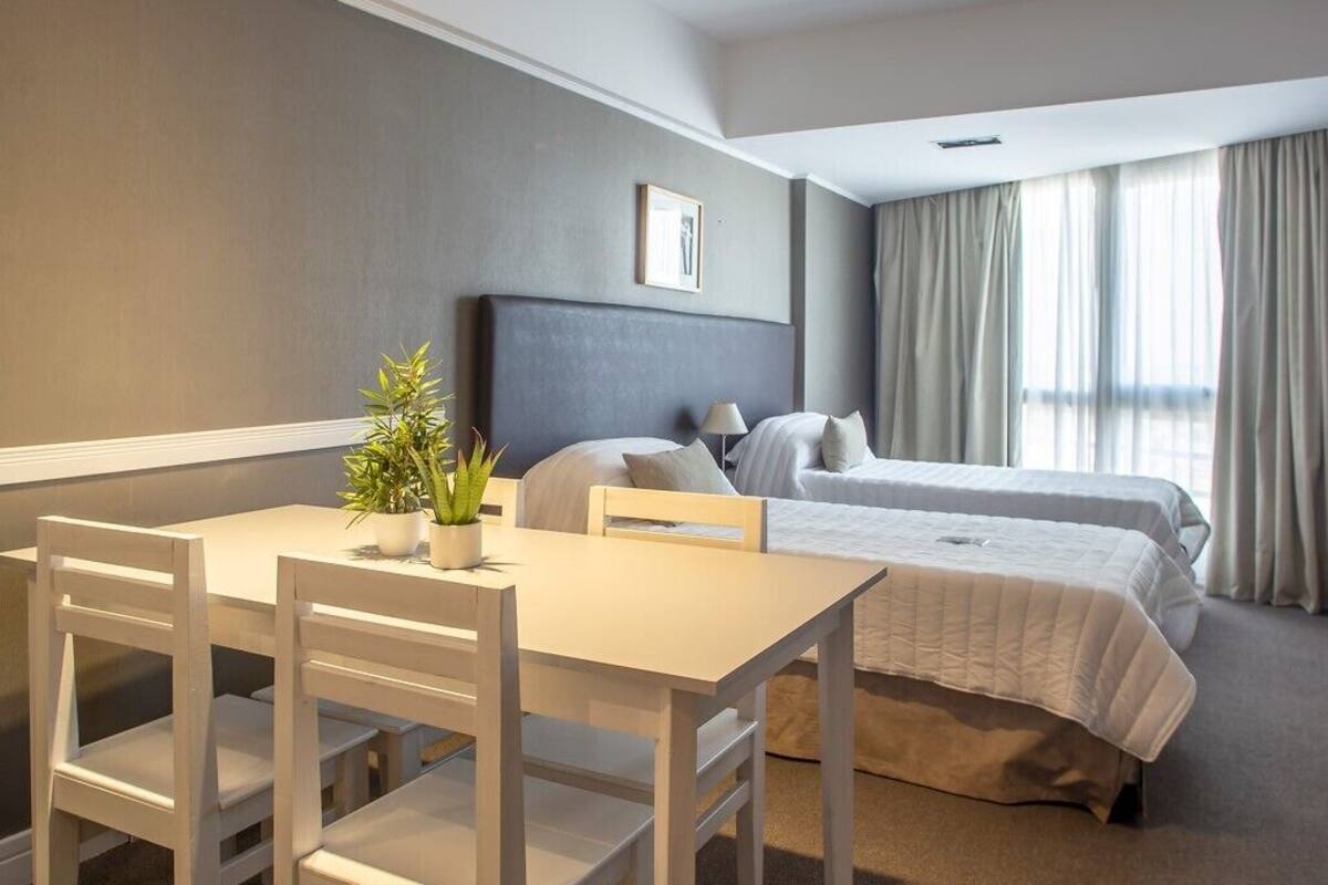 A hotel room with a bed, a desk with chairs, a potted plant, and a window with curtains.