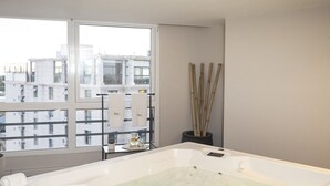 A large, rectangular hot tub with a white interior and a glass enclosure, positioned near a window with a city view. - Visit Hotel & Apart (San Luis)