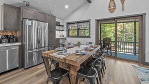 A modern kitchen with stainless steel appliances, a wooden dining table set for six, and a large sliding glass door leading to a deck with a view of trees.