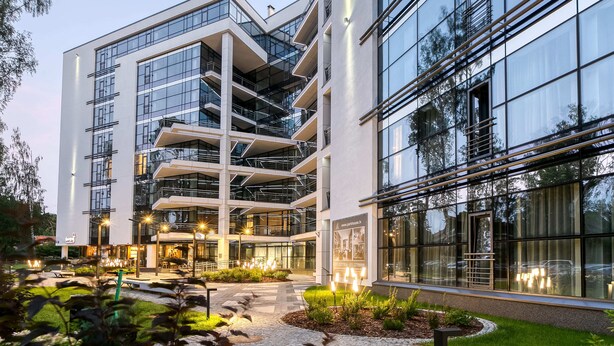 A modern multi-story building with a glass facade, outdoor seating area, and a landscaped garden. - Fresh and lovely apartment (Jurmala)