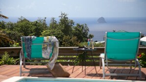 Área da piscina com duas cadeiras verdes, uma mesa pequena e vista para o oceano e uma ilha ao longe.