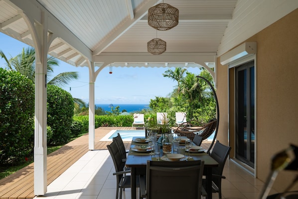 A covered outdoor dining area with a view of the ocean, a pool, and lush greenery.