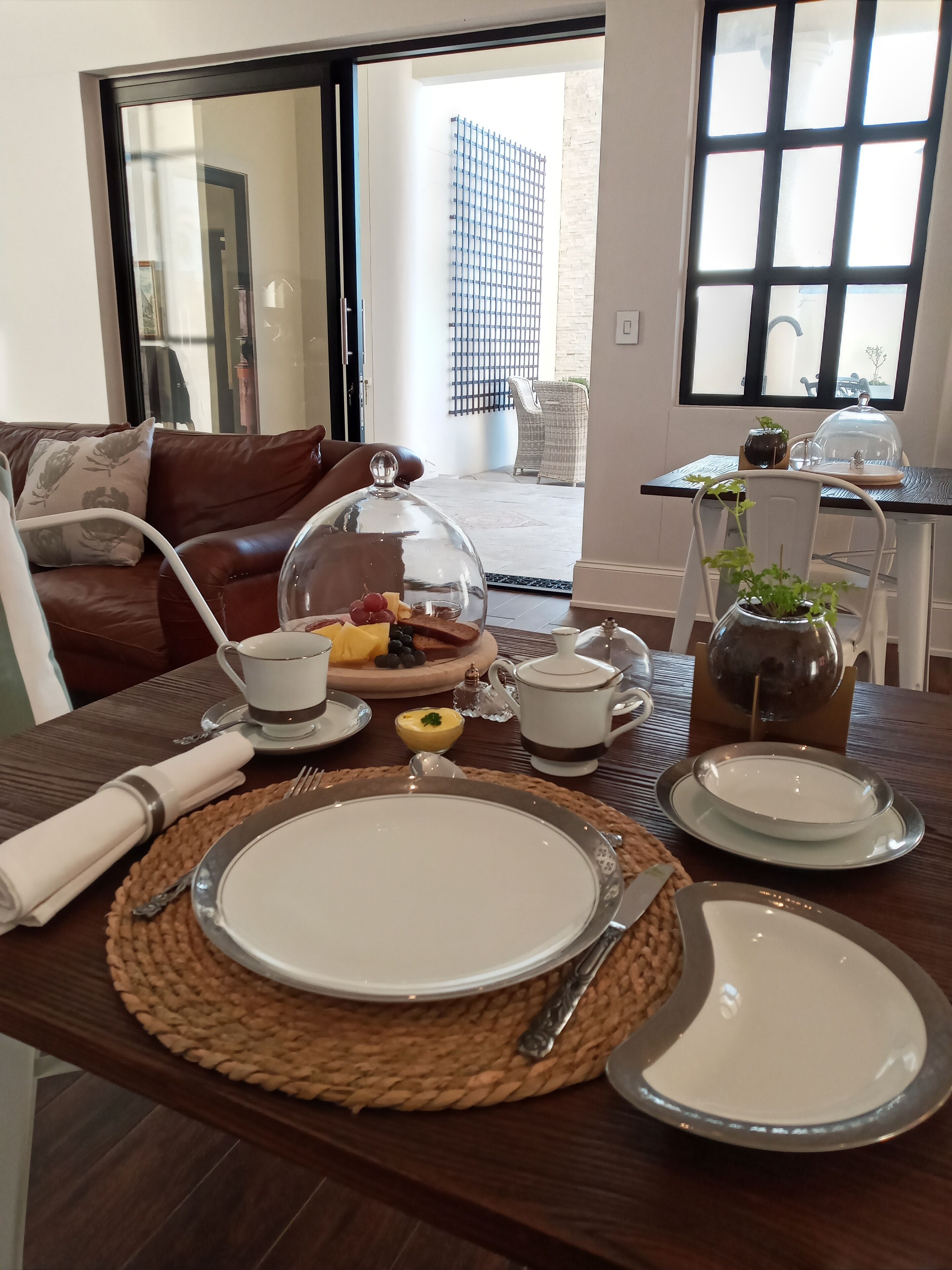 Un comedor con una mesa preparada para el desayuno, que incluye platos, cubiertos y una campana de cristal que cubre una disposición de frutas.