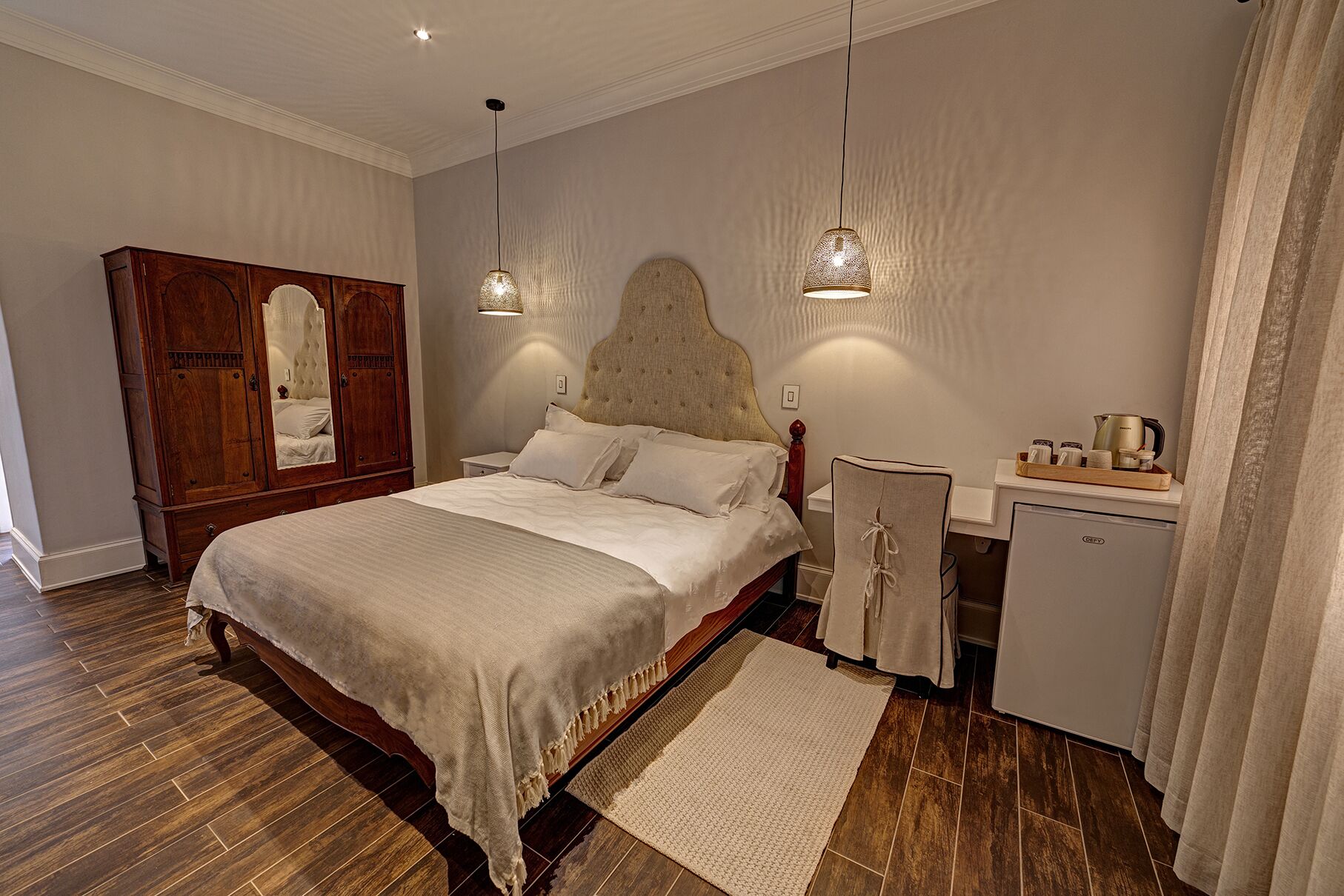 A bedroom with a large bed, a wardrobe, a nightstand, a desk with a kettle, and a refrigerator.
