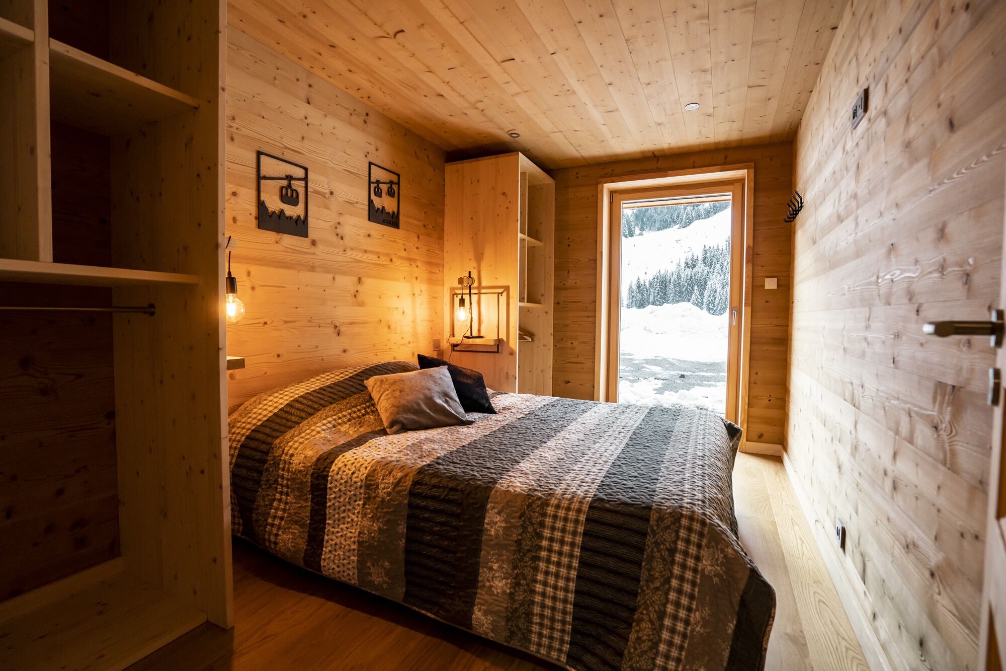 A cozy wooden cabin bedroom with a large bed, a wooden wardrobe, and a window offering a snowy mountain view.