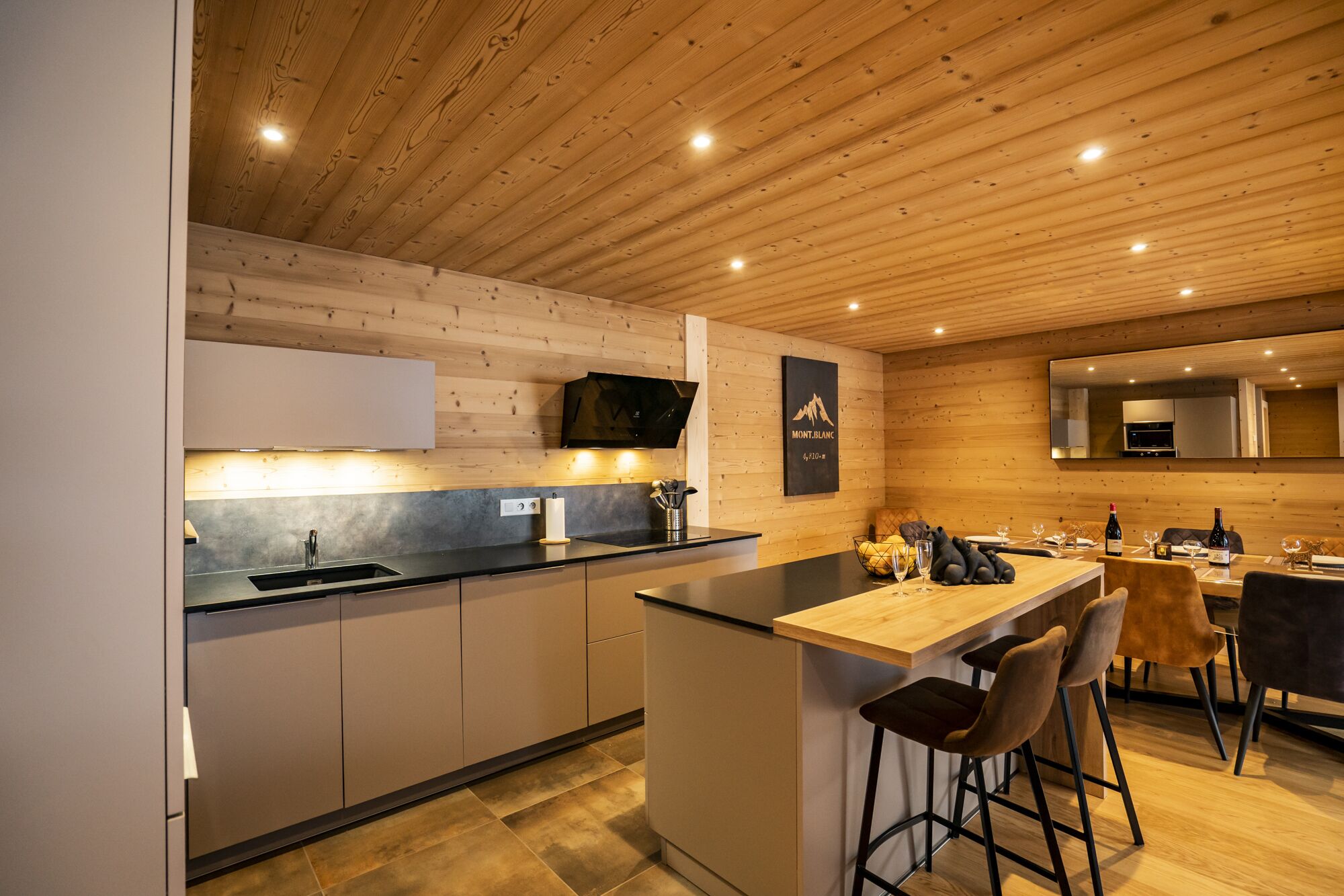A modern kitchen with wooden ceiling, a central island, bar stools, and a dining area with a table and chairs.