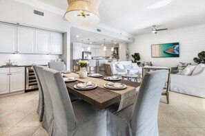 A modern kitchen with white cabinets, a dining area with a wooden table and grey upholstered chairs, and a living room with a white sofa and a large painting on the wall.