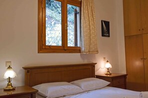 A bedroom with a wooden bed, two bedside lamps, a window with a view of trees, a wardrobe, and a framed picture on the wall.
