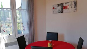 A dining area with a round table covered in a red tablecloth, surrounded by black chairs. There is a wall-mounted artwork above the table and a window with curtains to the left. - Les Demeures de la BeauClaire - Provençal Elegance, Pool, Air Conditioning, Warm Hospitality (Piolenc)