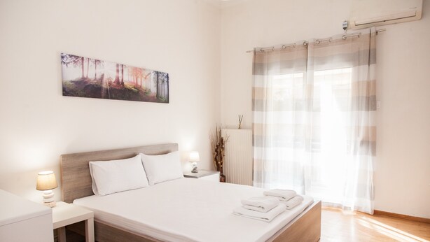 A modern hotel room with a large bed, a nightstand, a lamp, a painting on the wall, and a window with curtains. - Cozy apartment n Stavros Niarchos Park (Kallithea)