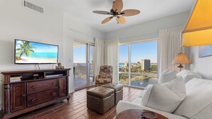 Ein geräumiges Wohnzimmer mit großem Fenster und Blick auf die Stadt, einem Deckenventilator, einem Fernseher und einer Holzkassette.