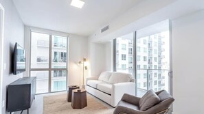 Een modern appartement met een witte bank, een televisie aan de muur en grote ramen met uitzicht op andere gebouwen.