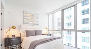 Een moderne slaapkamer met een groot raam, een bed met een witte hoofdbord, twee nachtkastjes met lampen, en een abstract kunstwerk aan de muur.