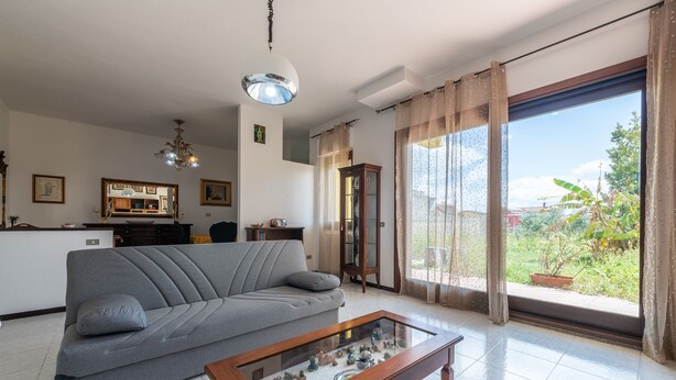A living room with a sofa, a coffee table, a chandelier, a large window with curtains, and a view of greenery outside.