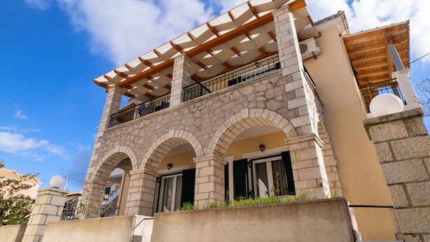 Un edificio con fachada de piedra, un balcón con pérgola de madera y ventanas arqueadas.
