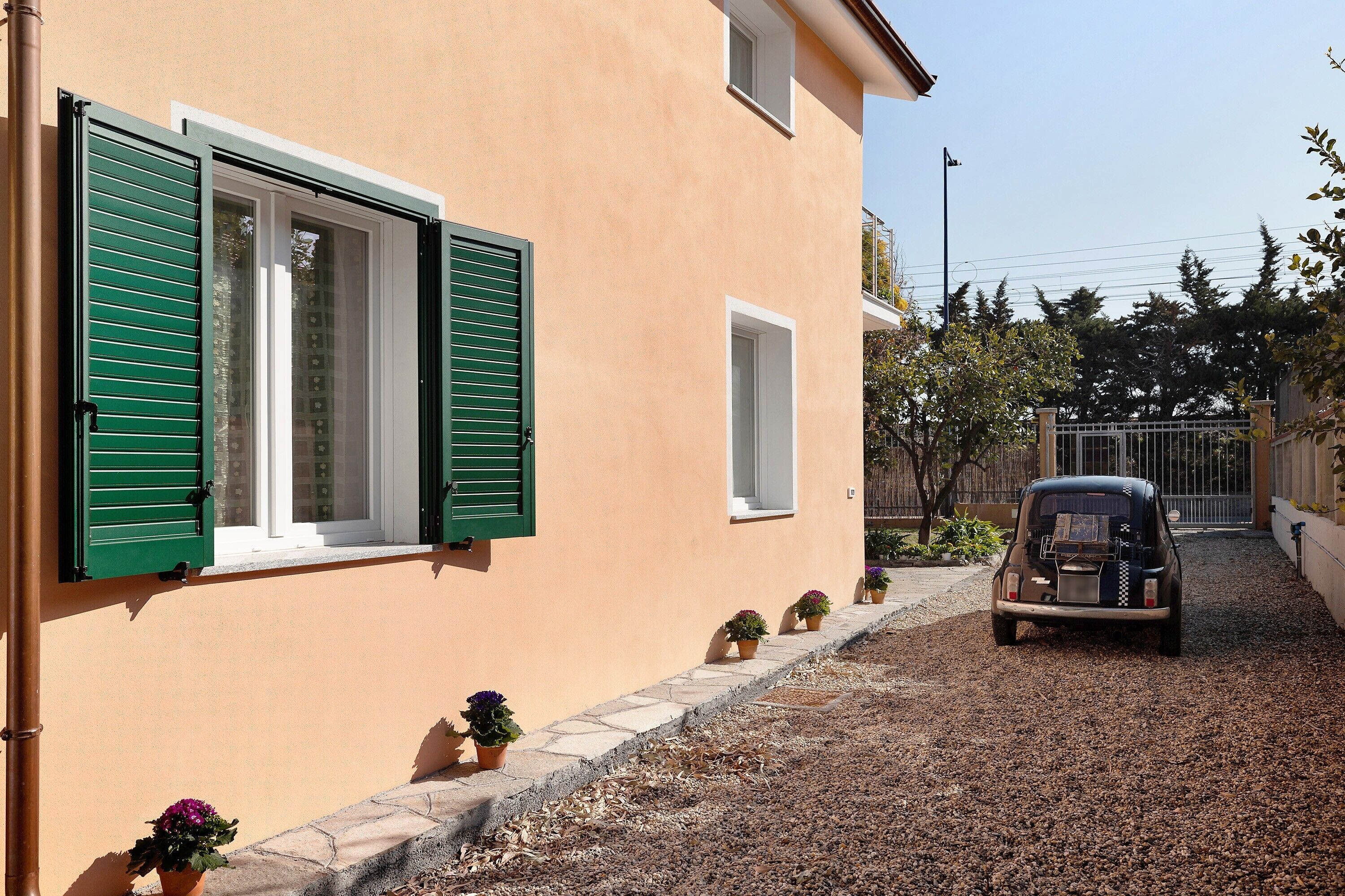 Un bâtiment à la façade beige, aux volets verts et à l’allée gravillonnée.