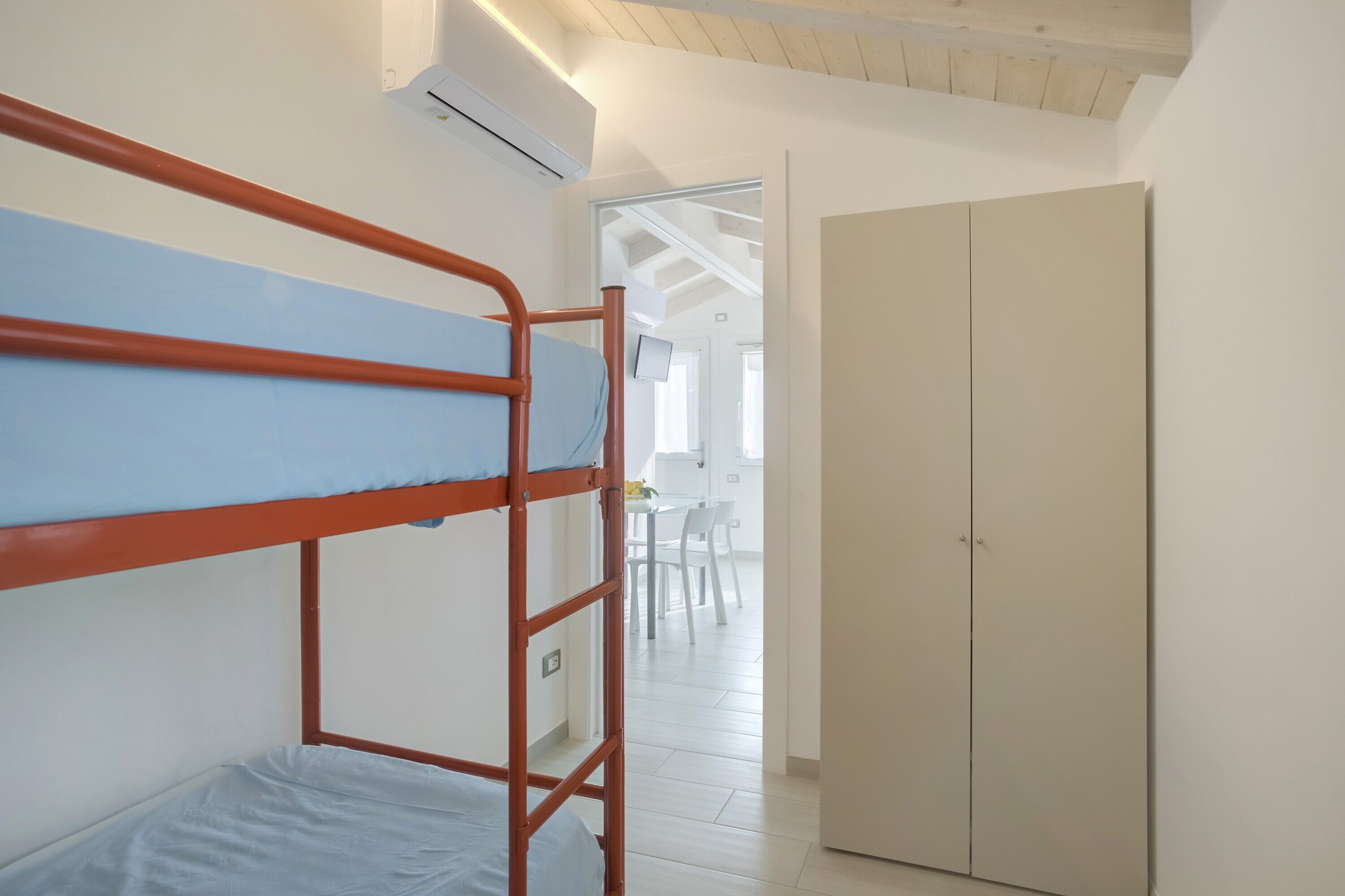 Ein Etagenbett mit blauer Matratze, ein Kleiderschrank und Blick in einen Raum mit Tisch und Stühlen.
