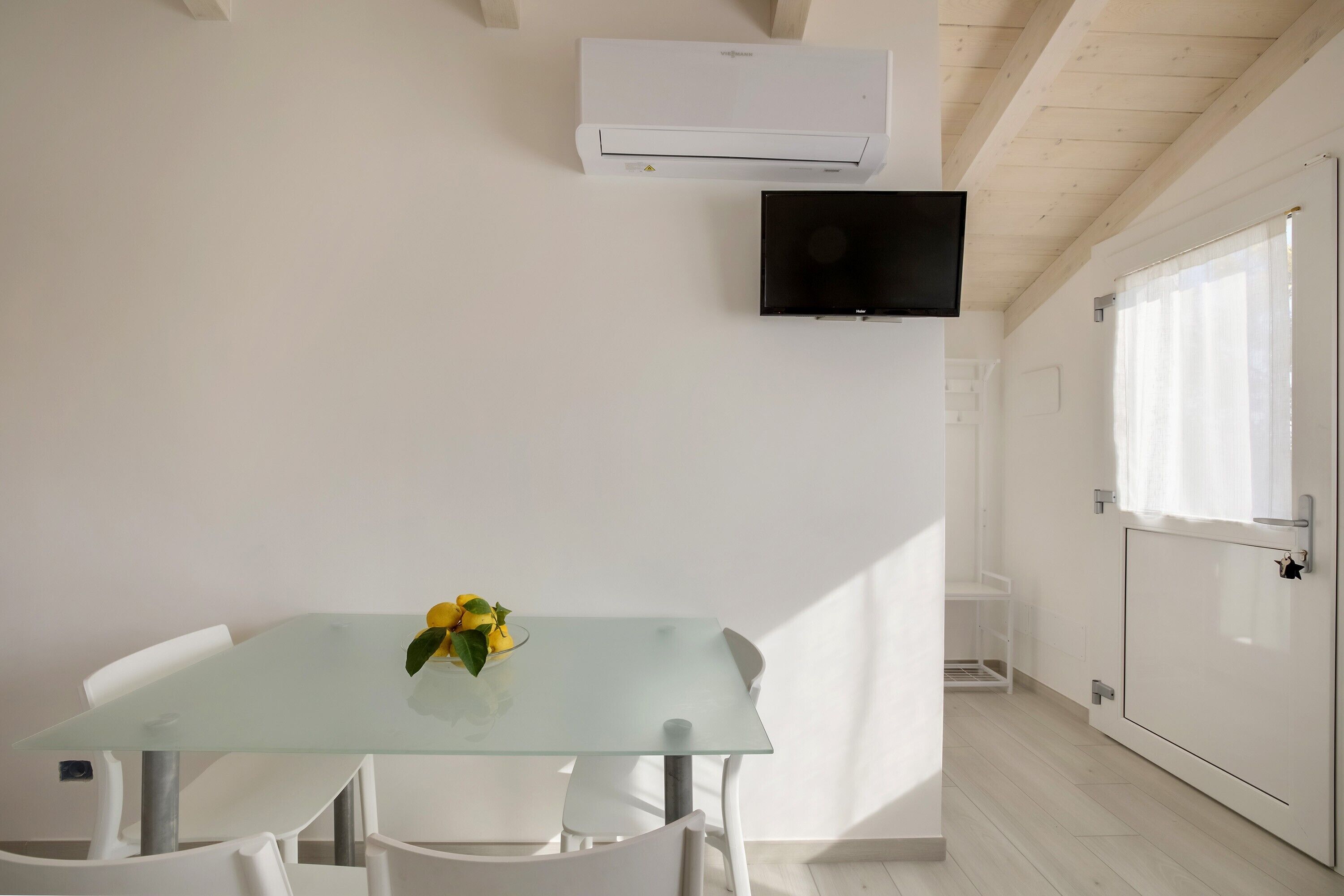 Quarto moderno com mesa de vidro, cadeiras brancas, televisão na parede e aparelho de ar condicionado.