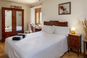 A neatly made bed with white linens, a wooden nightstand with a lamp, a wooden wardrobe, and a framed floral artwork on the wall.