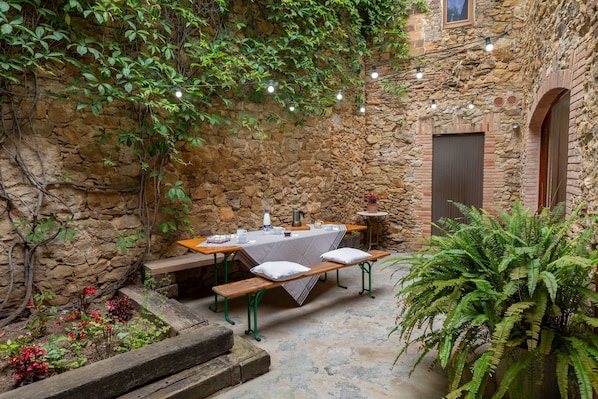 A stone-walled courtyard with a table and benches, string lights, and potted plants.