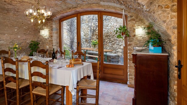 Une salle à manger rustique aux murs de pierre, meublée de meubles en bois et offrant une vue sur le paysage à travers une grande fenêtre en arc de cercle.