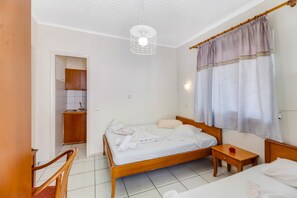 A hotel room with a wooden bed, a small bedside table, a chair, a wooden wardrobe, and a kitchen area.