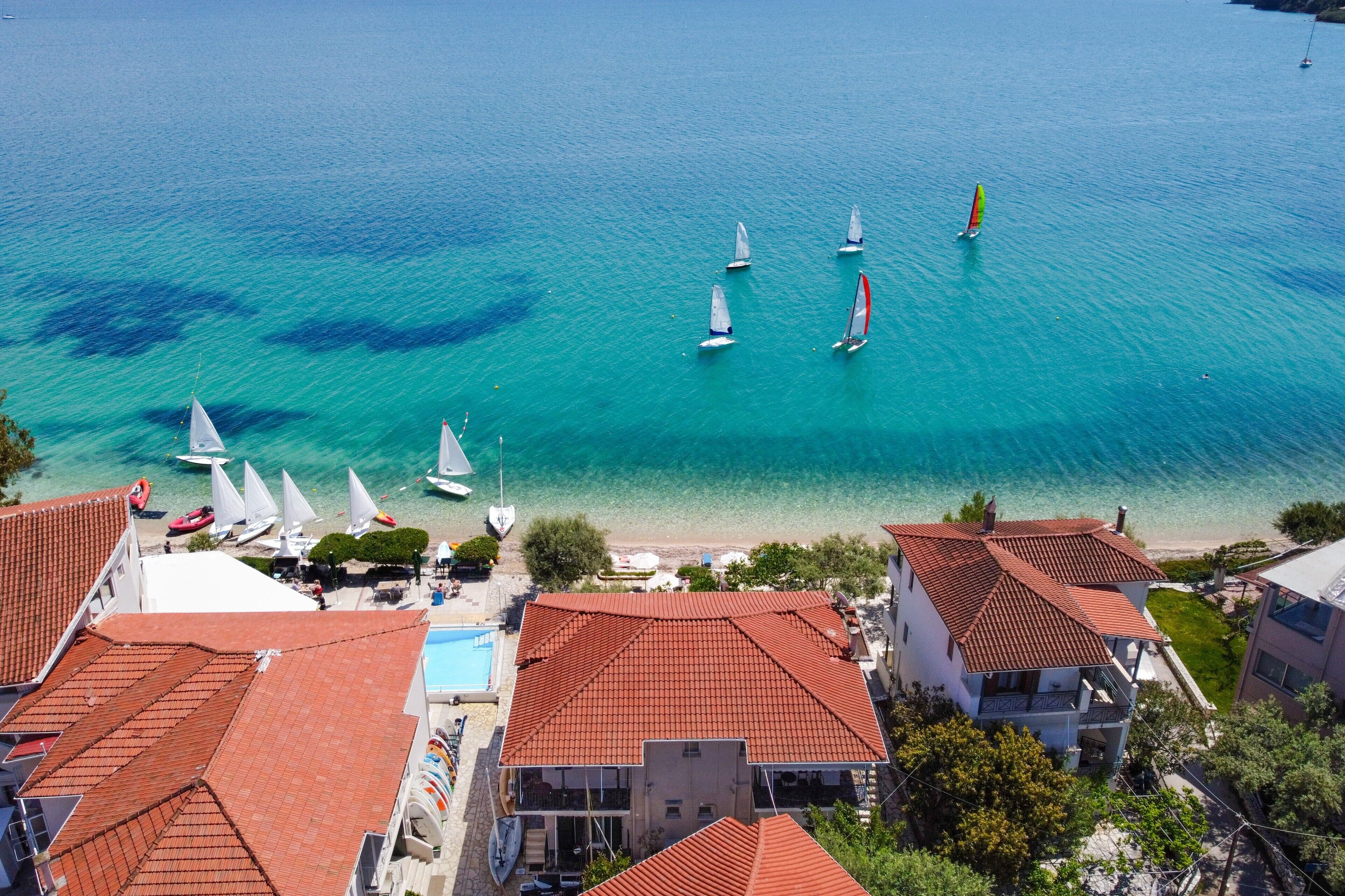 A coastal area with sailboats on turquoise water, buildings with red-tiled roofs, and a beach with umbrellas.
